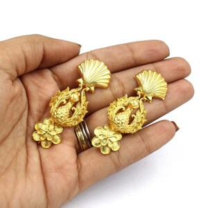 Puede incluir: Un par de pendientes de tono dorado con un diseño floral y un motivo de piña. Los pendientes son colgantes y tienen una parte superior en forma de concha.