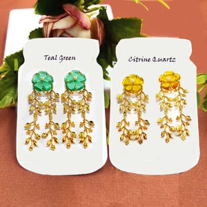 Puede incluir: Dos pares de pendientes colgantes con dise&ntilde;os de hojas doradas y piedras de vidrio en forma de flor. Los pendientes se muestran en tarjetas blancas con el texto "Teal Green" y "Citrine Quartz".