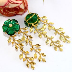 Puede incluir: Un par de pendientes colgantes de oro con detalles de flores de cristal verde y una perla. Los pendientes presentan un delicado dise&ntilde;o de hojas doradas.