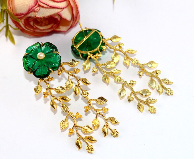 Puede incluir: Un par de pendientes colgantes de oro con detalles de flores de cristal verde y una perla. Los pendientes presentan un delicado dise&ntilde;o de hojas doradas.