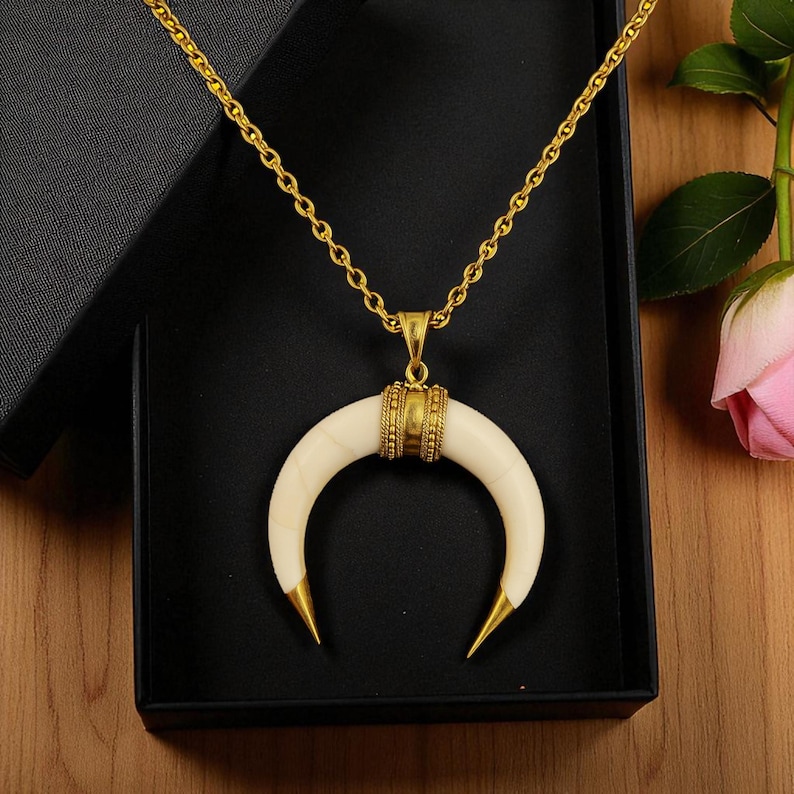 May include: A gold chain necklace featuring an ivory-coloured crescent moon pendant with gold-coloured tips and a gold band. The necklace is presented in a black box. The chain is a classic design.