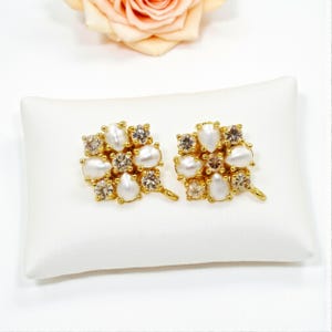 May include: A pair of gold-toned stud earrings. Each earring features a cluster of white, pear-shaped faux pearls and clear, round and square-cut crystals. The earrings are displayed on a white pillow.