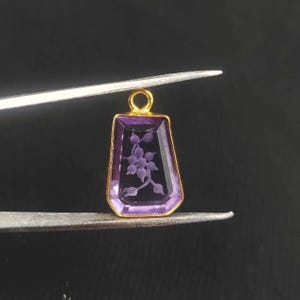 May include: A rectangular purple gemstone pendant with a gold-colored frame. The pendant features an etched floral design. The pendant is held by tweezers against a dark background. Jewelry.