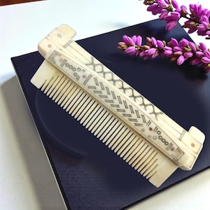 May include: Ivory-colored folding comb with intricate black and red geometric designs. The comb is open, revealing fine teeth. A sprig of purple flowers rests nearby, set against a dark blue surface.