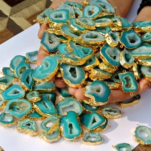 Op de afbeelding: Een grote stapel teal agaat plakjes met gouden randen. De agaat plakjes hebben een natuurlijke, onregelmatige vorm en een gat geboord aan de bovenkant. De plakjes zijn perfect voor het maken van sieraden of andere knutselprojecten.