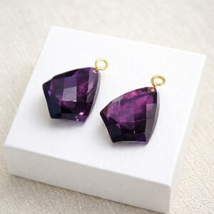 May include: Two faceted, deep purple amethyst-colored pendants with irregular, geometric shapes. Each pendant features a small, circular gold-toned metal jump ring attached to the top, allowing for easy suspension on a necklace or earring wire.