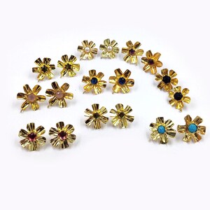 May include: A collection of gold-toned metal flower-shaped earring components arranged in pairs. Each piece features a textured, petal-like design with a central setting containing various gemstones, including pearls, blue, pink, and turquoise stones.