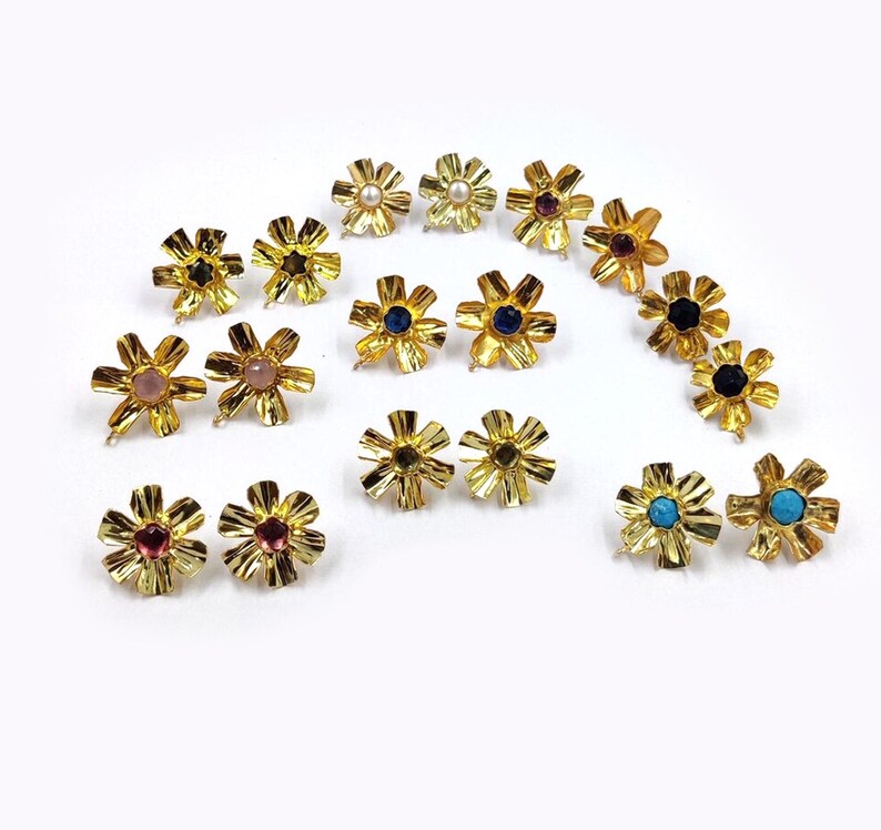 May include: A collection of gold-toned metal flower-shaped earring components arranged in pairs. Each piece features a textured, petal-like design with a central setting containing various gemstones, including pearls, blue, pink, and turquoise stones.