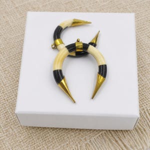 Peut inclure: Deux pendentifs en forme de croissant avec des segments altern&eacute;s noirs et cr&egrave;me. Chaque extr&eacute;mit&eacute; des pi&egrave;ces incurv&eacute;es est surmont&eacute;e d'une pointe en m&eacute;tal dor&eacute;, et une petite boucle en m&eacute;tal est fix&eacute;e au centre pour permettre la suspension.