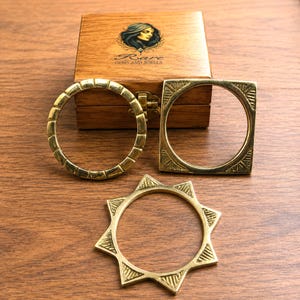 Può includere: Tre braccialetti in ottone con diversi design geometrici sono esposti su una superficie di legno. Uno è rotondo con un motivo segmentato, un altro è quadrato con un centro circolare e il terzo è a forma di stella. Una scatola di legno con la scritta "Rare Gems and Jewels" è sullo sfondo.