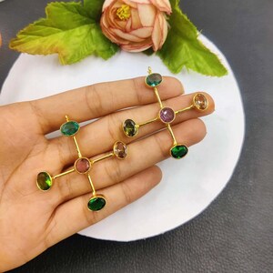 Puede incluir: Dos collares con colgantes dorados, cada uno con un dise&ntilde;o en forma de cruz con piedras preciosas de colores. Las piedras preciosas ovaladas incluyen tonos verdes, rosas y marrones. Los colgantes se muestran sobre una superficie blanca.