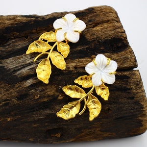 May include: A pair of floral earrings featuring white flower-shaped pendants with gold-colored accents and leaves. The earrings are set against a dark wooden surface, creating a contrast that highlights the jewelry's design.