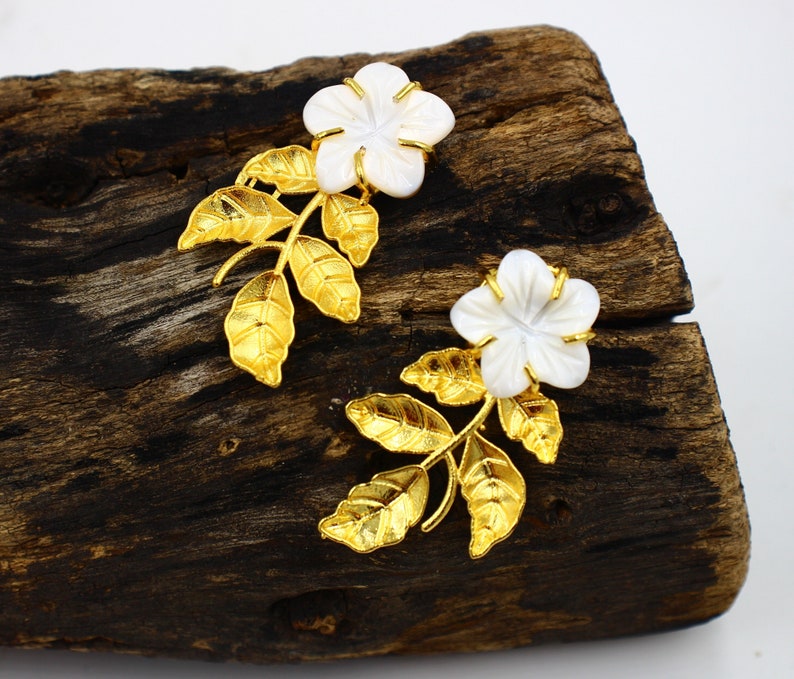 May include: A pair of floral earrings featuring white flower-shaped pendants with gold-colored accents and leaves. The earrings are set against a dark wooden surface, creating a contrast that highlights the jewelry's design.