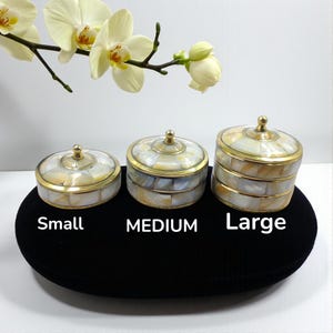 Könnte beinhalten: Drei dekorative runde Boxen in den Größen klein, mittel und groß. Die Boxen haben ein Perlmutt-Design mit goldfarbenem Rand und einen kleinen Knopf auf dem Deckel. Der Text "Small", "Medium" und "Large" steht darunter.