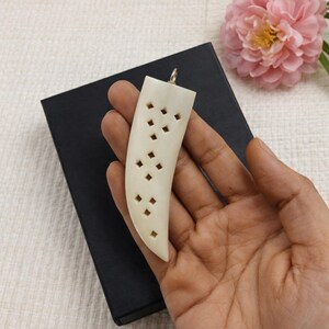 May include: Ivory-colored pendant with a curved, elongated shape and a series of diamond-shaped cutouts. The pendant has a silver-colored loop at the top for hanging. The pendant is held in a hand, with a black box in the background.