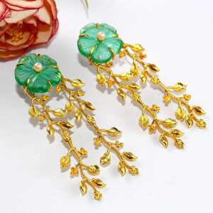 Puede incluir: Un par de pendientes colgantes de oro con detalles de cristal verde en forma de flor y una peque&ntilde;a perla en el centro de cada flor. Los pendientes presentan delicadas ramas doradas con hojas.