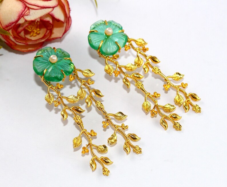 Puede incluir: Un par de pendientes colgantes de oro con detalles de cristal verde en forma de flor y una peque&ntilde;a perla en el centro de cada flor. Los pendientes presentan delicadas ramas doradas con hojas.