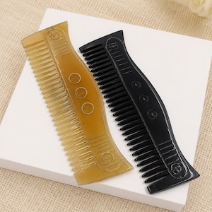May include: Two hair combs, one in a translucent amber tone and one in solid black, featuring a curved spine with three circular indentations in the center and horizontal line detailing at the top and bottom ends of the handle area.
