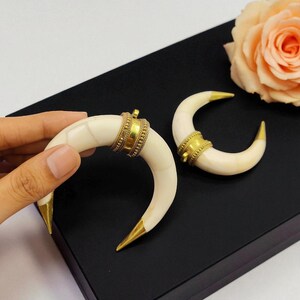 May include: A pair of ivory-coloured, crescent-shaped earrings with gold-coloured tips and decorative gold bands. One earring is held by a hand. The earrings are on a black surface, with a peach rose in the background. The earrings have a unique, curved design.