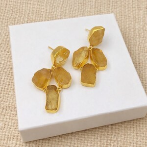 May include: Gold-toned earrings featuring clusters of rough-cut, amber-colored gemstones. The earrings have a post-back design and are displayed on a white surface. The gemstones have a natural, irregular shape, adding a unique touch to the jewelry.