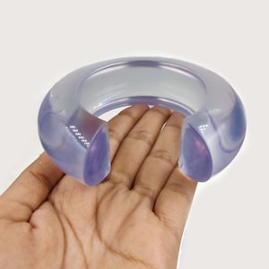 Peut inclure: Un bracelet rigide en résine translucide de couleur violette. Le bracelet est épais et de forme arrondie.