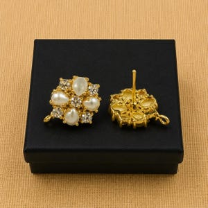 May include: A pair of gold-colored stud earrings. Each earring features a cluster of white, pear-shaped faux pearls and clear, round crystals. The earrings are displayed on a black box.