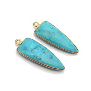 May include: Two turquoise-colored, teardrop-shaped pendants with gold-colored metal trim. Each pendant has a small loop at the top for attaching to a chain or other jewelry component. The pendants are set against a white background.