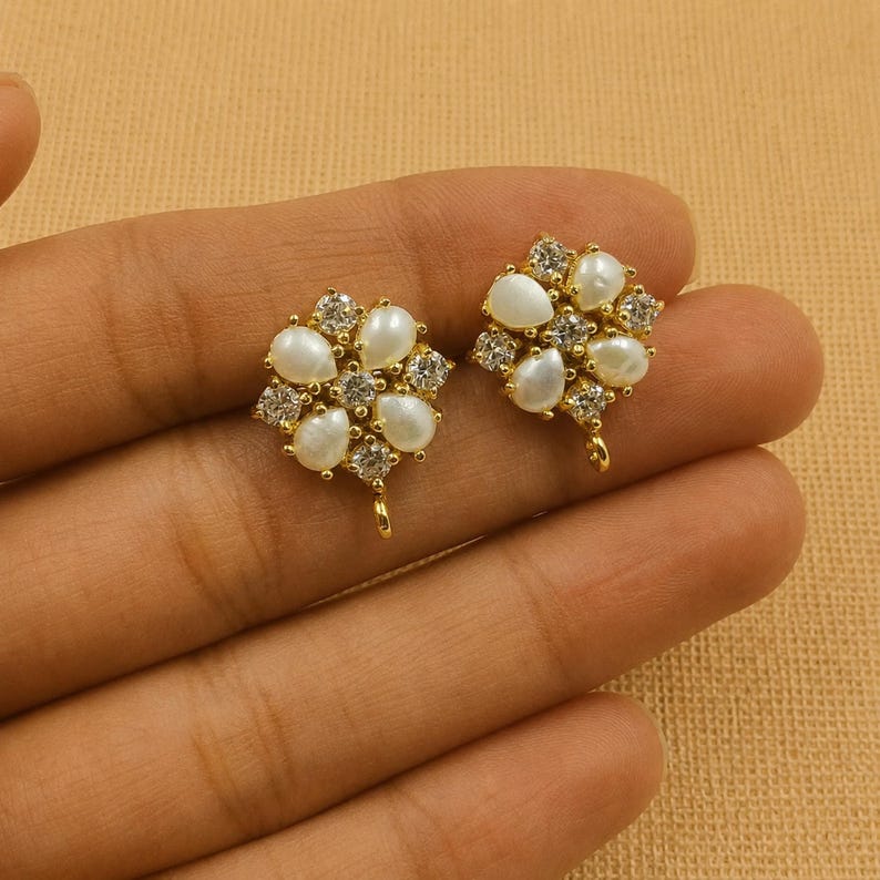 May include: Two gold-toned stud earrings, each featuring a floral design. Each earring has a cluster of white, teardrop-shaped pearls and clear, sparkling crystals. The earrings have a small loop at the bottom for attachment.
