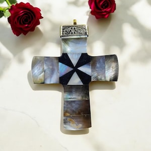 Può includere: Un grande pendente a forma di croce ornato con intarsi in madreperla. La croce presenta una parte superiore in metallo color argento con incisioni decorative e un design geometrico in bianco e nero al centro. Due rose rosse sullo sfondo.