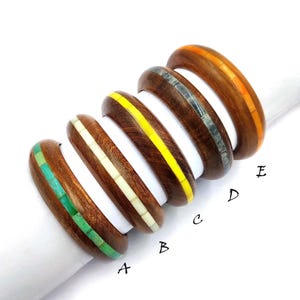 May include: Five wooden bangles with inlaid colourful designs. The bangles are stacked on top of each other, with the top bangle having an orange inlay, the next bangle having a grey inlay, the next bangle having a yellow inlay, the next bangle having a white inlay, and the bottom bangle having a green inlay. The bangles are labelled A through E.