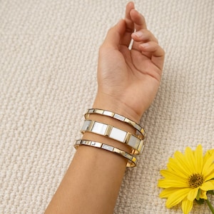 Op de afbeelding: Vier goudkleurige armbanden met rechthoekige witte en gouden accenten zijn op een pols gestapeld. De armbanden worden gedragen op een lichtgekleurde arm. Een gele bloem met een groene stengel staat in de rechter benedenhoek. De achtergrond is een lichtbeige getextureerd oppervlak.
