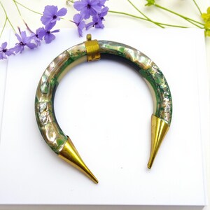 Può includere: Un braccialetto curvo e aperto con un design a conchiglia verde e iridescente. Il braccialetto ha dettagli in metallo color oro alle estremità e una chiusura. Un gioiello unico.