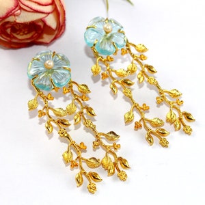 Puede incluir: Un par de pendientes de oro con un dise&ntilde;o de flor azul claro y un acento de perla. Los pendientes presentan un delicado dise&ntilde;o de hojas que cuelga de la flor.