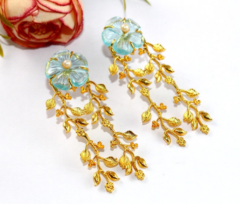 Puede incluir: Un par de pendientes de oro con un dise&ntilde;o de flor azul claro y un acento de perla. Los pendientes presentan un delicado dise&ntilde;o de hojas que cuelga de la flor.