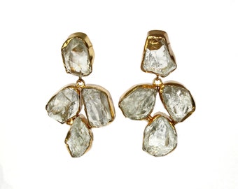 May include: A pair of drop earrings featuring raw, clear, irregular-shaped stones set in gold-toned metal. Each earring consists of a single top stone connected to a cluster of three stones below, creating a dangling, multi-stone design.