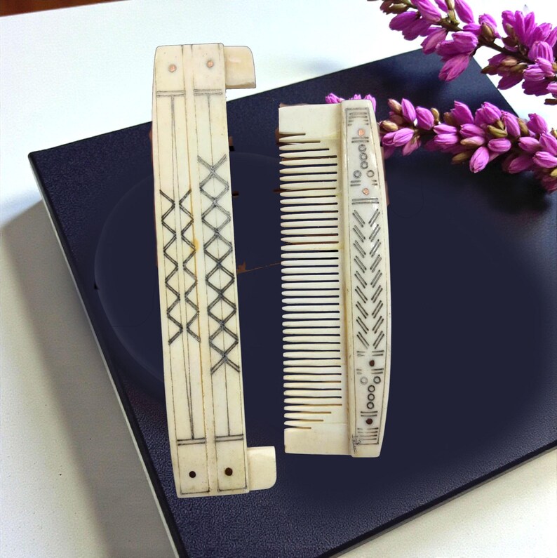 May include: Two antique ivory-colored combs with intricate black etched designs. One comb is foldable, featuring a zigzag pattern, while the other has a row of teeth and a decorative border. A sprig of purple flowers rests nearby.