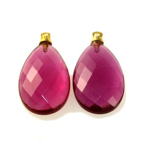 May include: Two faceted, teardrop-shaped, magenta-colored gemstone pendants with gold-colored caps. The pendants are displayed against a white background. The gemstones have a geometric cut, reflecting light.