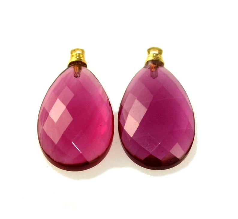 May include: Two faceted, teardrop-shaped, magenta-colored gemstone pendants with gold-colored caps. The pendants are displayed against a white background. The gemstones have a geometric cut, reflecting light.