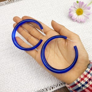 Puede incluir: Un par de pendientes de aro grandes y circulares hechos de material acrílico azul profundo y translúcido. Los pendientes presentan un perfil liso y redondeado e incluyen finos postes de metal plateado para su sujeción, con un diseño vibrante.