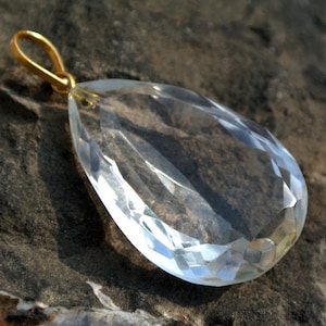 May include: A clear, faceted crystal pendant with a gold-colored bail. The teardrop-shaped gemstone is highly polished, reflecting light and showcasing its intricate cuts. The pendant is displayed against a textured, dark gray surface.