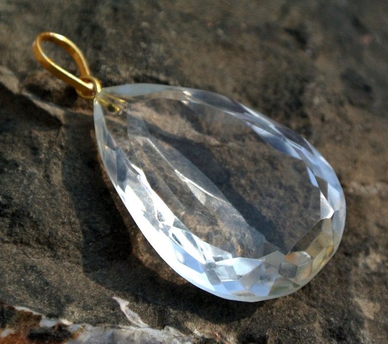 May include: A clear, faceted crystal pendant with a gold-colored bail. The teardrop-shaped gemstone is highly polished, reflecting light and showcasing its intricate cuts. The pendant is displayed against a textured, dark gray surface.