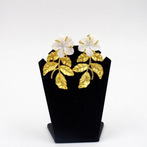 May include: A pair of floral earrings. Each earring features a white flower with gold-colored accents, set atop a gold-colored stem with detailed leaves. The earrings are displayed on a black stand against a white background.