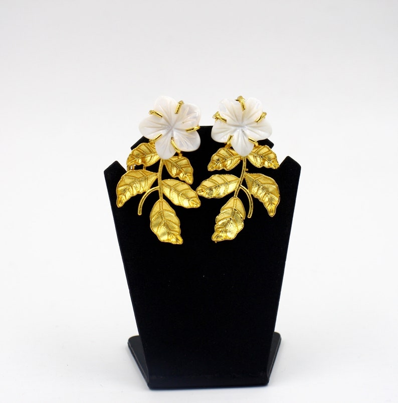 May include: A pair of floral earrings. Each earring features a white flower with gold-colored accents, set atop a gold-colored stem with detailed leaves. The earrings are displayed on a black stand against a white background.
