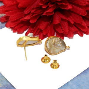 May include: A pair of gold-tone earrings with a white and iridescent shell design. The earrings are shown on a white surface with two extra earring backs.