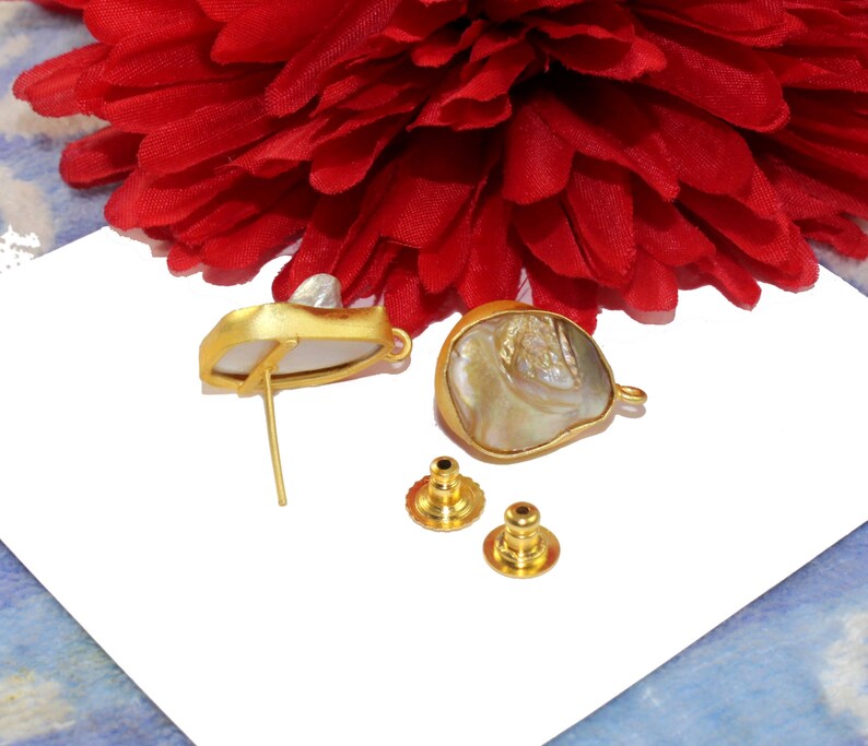 May include: A pair of gold-tone earrings with a white and iridescent shell design. The earrings are shown on a white surface with two extra earring backs.