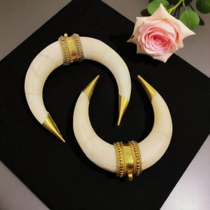 May include: Two ivory-coloured, crescent-shaped bangles with gold-coloured tips and decorative gold bands. The bangles are arranged on a black surface, with a pink rose in the background. The jewellery has a vintage or tribal aesthetic.