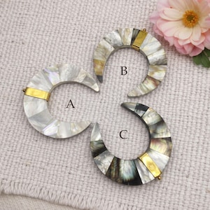 May include: Three crescent-shaped pendants with iridescent mother-of-pearl segments. Each pendant has a gold-colored metal accent. The pendants are labeled A, B, and C. A pink flower is in the background.