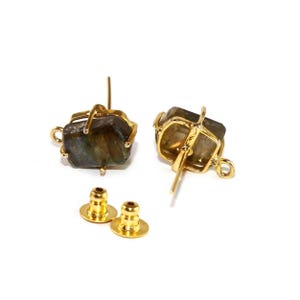 May include: A pair of gold-colored stud earrings with rough-cut, iridescent, gray-green stones. The stones are held in place by a delicate gold wire frame. Two gold earring backs are also visible.