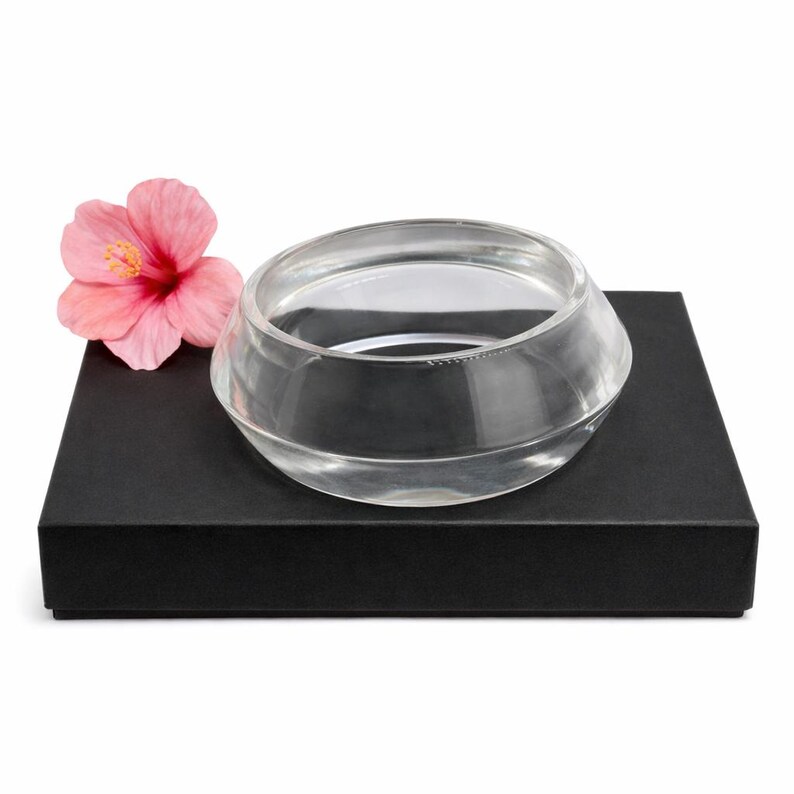 May include: A clear, round glass bowl with a unique, angled design sits atop a black box. A pink hibiscus flower with a yellow center rests beside the bowl. The bowl is likely for decorative purposes.