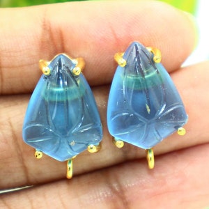 May include: A pair of blue teardrop-shaped earrings with gold accents. The earrings have a carved floral design and are attached to a gold hook.